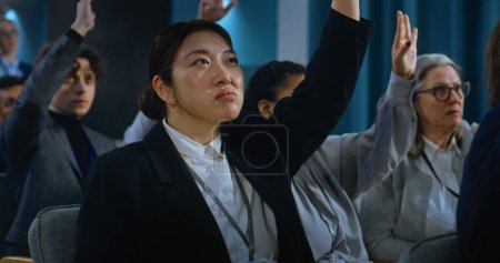 Diverse press workers sit in the conference hall raising hands. Asian female journalist asks question, listens the answer of organization representative or politician during press campaign for media.