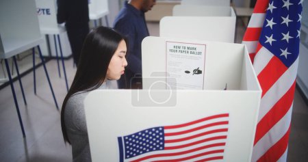 Foto de Mujer asiática viene a la cabina de votación en el centro de votación, toma decisiones y llena el boletín de papel. Ciudadano estadounidense durante el Día Nacional de Elecciones en los Estados Unidos de América. Deber cívico y patriotismo. - Imagen libre de derechos