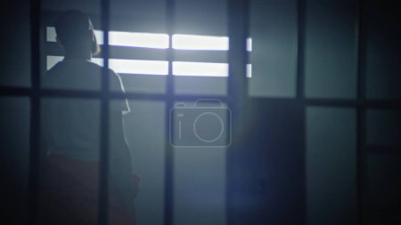 Un prisionero de uniforme naranja camina en la celda de la prisión, luego se sienta en la cama y comienza a leer la Biblia. Recluso religioso cumple condena de prisión en la cárcel. Centro de detención o correccional.