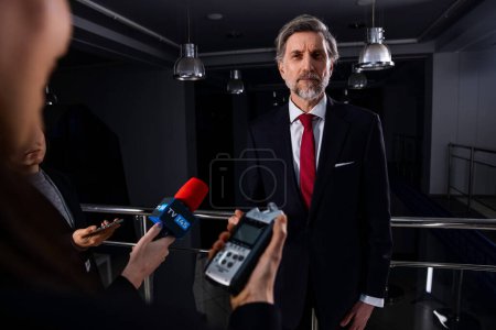 Confident and serious male diplomat or businessman talks with news journalists and gives interview for media and television in government building. Political speech of politician at press conference.