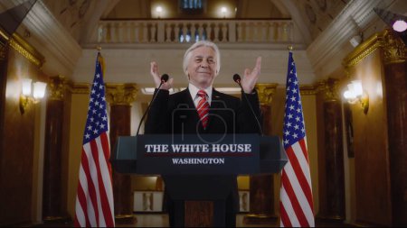 Photo for Joyful senior politician poses on cameras for media and television. New President of the United States smiles and gestures during press conference in the White House. American flags in the background. - Royalty Free Image