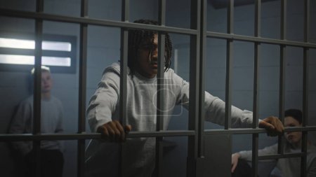Foto de Adolescentes multiétnicos tras las rejas de metal en la celda de la prisión miran a la cámara. Jóvenes prisioneros, criminales cumplen penas de prisión por crímenes en la cárcel. Centro de detención juvenil o correccional. Retrato - Imagen libre de derechos