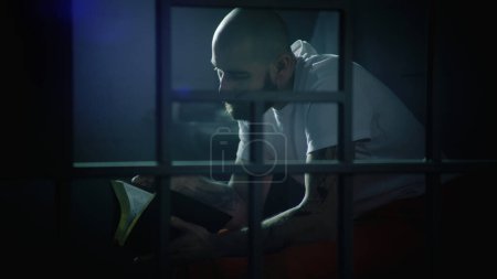 Male prisoner in orange uniform sits on bed in prison cell, reads Bible. Illegally convicted man serves imprisonment term in jail. View through metal bars. Detention center or correctional facility.