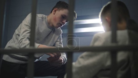 Téléchargez les photos : Un adolescent caucasien joue aux cartes en cellule avec d'autres détenus. Les jeunes criminels purgent une peine d'emprisonnement. Centre de détention pour mineurs ou établissement correctionnel. Vue à travers des barres métalliques. - en image libre de droit