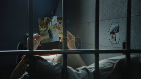 Male prisoner lies on bed in prison cell, looks at pictures of family. Inmate or criminal serves imprisonment term for crime in jail. View through metal bars. Detention center or correctional facility