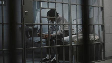 African American young prisoner eats disgusting prison food from iron bowl sitting on bed in prison cell. Teenage criminal, inmate serves imprisonment term for crime in jail. Youth detention center.
