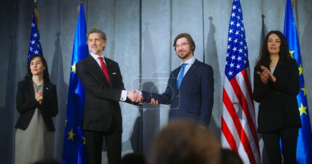 Photo for European Union representative and mature president of the United States together shake hands after press conference. World leaders during meeting with journalists. Backdrop with American and EU flags. - Royalty Free Image
