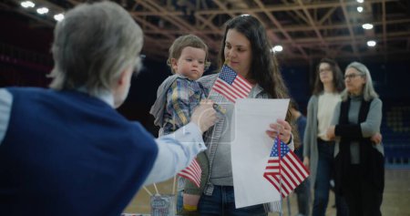 Photo for Woman with baby on hands talks with polling officer and takes bulletin. Female American citizen comes to vote in polling station. Political races of US presidential candidates. National Election Day. - Royalty Free Image