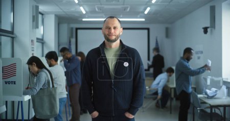 Téléchargez les photos : Portrait d'un homme caucasien, électeur des États-Unis d'Amérique. L'homme barbu se tient dans un bureau de vote moderne, pose, sourit, regarde la caméra. Contexte avec isoloirs. Concept du devoir civique. - en image libre de droit