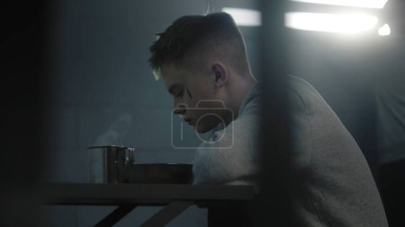 Caucasian teenage prisoner eats prison food at the table in prison cell. Young criminal serves term for crime in youth detention center or jail. Another inmate looks at the window in the background.