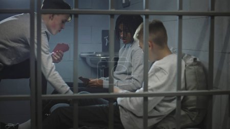 Foto de Tres diversos adolescentes juegan juegos de cartas en la celda de la prisión. Jóvenes prisioneros, criminales cumplen pena de prisión por crímenes en la cárcel. Centro de detención juvenil o correccional. Vista a través de barras de metal. - Imagen libre de derechos