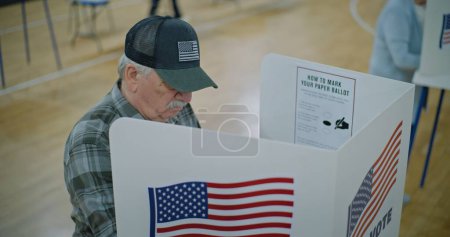 Elderly male voter with bulletin in hands comes to voting booth. American citizens come to vote in polling station. Political races of US presidential candidates. National Election Day. Dolly shot.
