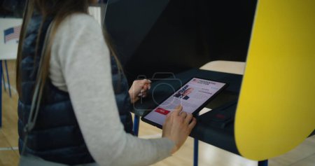 US-Bürger nutzen Tablet-Computer zur Stimmabgabe. Kaukasische Frau, denkt eine Wählerin und trifft die Wahl, als sie in der Nähe der Wahlkabine im Wahllokal steht. Präsidentschaftswahl in den Vereinigten Staaten von Amerika.