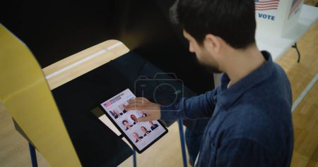 Kaukasier trifft Wahl und wählt US-Präsidentschaftskandidat in Wahlkabine per Tablet-Computer. US-Bürger im Wahllokal während des National Election Day in den Vereinigten Staaten. Hoher Winkel.