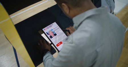 Foto de ZAPORIZHYA, UCRANIA -15 DE MARZO DE 2024: El hombre afroamericano, el votante masculino, elige y vota por Donald Trump en la cabina de votación usando una tableta. Ciudadano estadounidense durante las elecciones presidenciales en el - Imagen libre de derechos