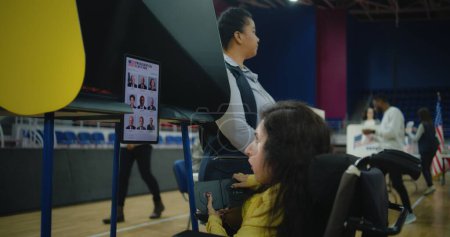 Wahlhelferin berät Frau mit körperlicher Behinderung Eine US-Wählerin mit SMA nutzt in der Wahlkabine im Wahllokal ferngesteuerte Knöpfe und wählt am Tablet-Computer. Wahltag in den Vereinigten Staaten.