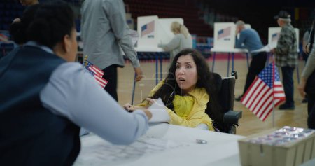 Woman with SMA in motorized wheelchair comes and talks with African American polling officer. Political races of US presidential candidates in polling station. National Election Day in United States.