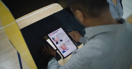 Foto de ZAPORIZHYA, UCRANIA -15 DE MARZO DE 2024: El votante afroamericano toma la decisión de Joe Biden y Donald Trump en la cabina de votación usando una tableta. Ciudadano estadounidense durante las elecciones presidenciales en Estados Unidos - Imagen libre de derechos