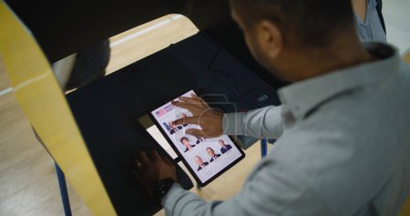 Afroamerikaner, männliche Wähler wählen den US-Präsidentschaftskandidaten in der Wahlkabine per Tablet-Computer. US-Bürger im Wahllokal. Wahltag in den USA. Hoher Winkel.
