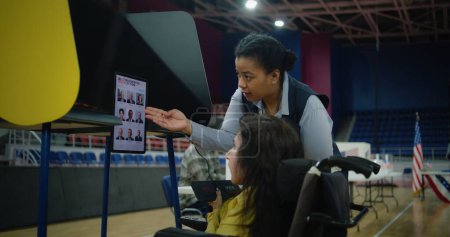 Wahlhelferin berät Frau mit körperlicher Behinderung Eine US-Wählerin mit SMA nutzt in der Wahlkabine im Wahllokal ferngesteuerte Knöpfe und wählt am Tablet-Computer. Wahltag in den Vereinigten Staaten.