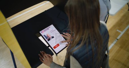 Kaukasierin trifft Wahl und wählt US-Präsidentschaftskandidatin in Wahlkabine mit Tablet-Computer US-Bürger im Wahllokal am Nationalwahltag in den Vereinigten Staaten. Hoher Winkel.