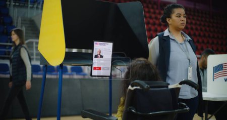 Foto de Votante mujer, mujer con SMA en silla de ruedas vota en cabina de votación en el centro de votación. Ciudadano de los Estados Unidos con discapacidad utiliza tableta para votar. Elecciones presidenciales en Estados Unidos. - Imagen libre de derechos