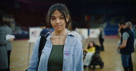 Glückliche junge US-Bürgerin nach ihrer Stimmabgabe bei den Präsidentschaftswahlen in den Vereinigten Staaten. Afroamerikanerin mit Abzeichen lächelt und blickt in die Kamera, die im Wahllokal steht. Bürgerpflicht. Porträt.
