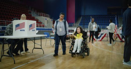 Téléchargez les photos : Une femme avec SMA en fauteuil roulant électrique vient avec le préposé au scrutin à l'isoloir. La femme afro-américaine donne le contrôle de vote à l'électeur. Journée nationale des élections aux États-Unis. Notion de devoir civique. - en image libre de droit