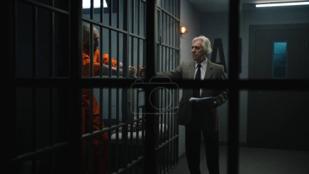 Foto de Criminal en uniforme naranja se apoya en las barras de celdas de la prisión, habla con el abogado y lee el contrato de abogado. El recluso cumple condena de prisión por delito en correccional. Gángster en centro de detención. - Imagen libre de derechos
