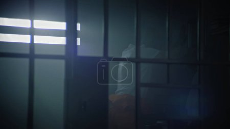 Männlicher Häftling in orangefarbener Uniform geht in der Gefängniszelle umher, setzt sich dann auf das Bett und beginnt, Bibel zu lesen. Religiöser Häftling verbüßt Haftstrafe im Gefängnis. Haftanstalt oder Justizvollzugsanstalt.