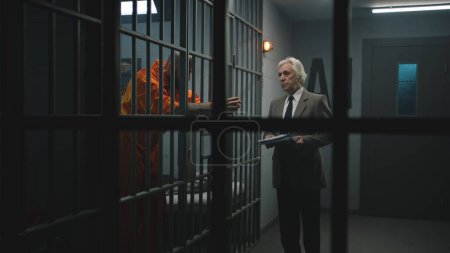 Foto de Criminal en uniforme naranja se apoya en las barras de celdas de la prisión, habla con el abogado y lee el contrato de abogado. El recluso cumple condena de prisión por delito en correccional. Gángster en centro de detención. - Imagen libre de derechos