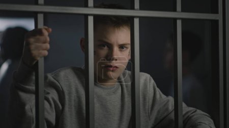 Caucasian teenager with face tattoos stands in prison cell in jail, holds metal bars and looks at camera. Diverse young inmates talk in the background. Juvenile detention center or correctional