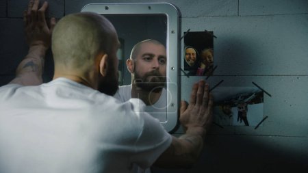 Depressed male prisoner stands in prison cell, looks at the pictures with family hanging near the mirror. Inmate serves imprisonment term for crime in jail. Detention center or correctional facility.