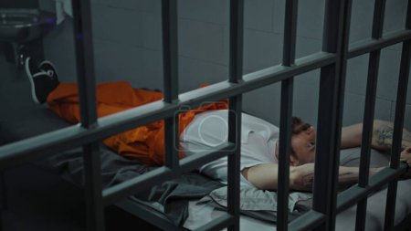 Photo for Inmate in orange uniform lies on prison cell bed. Prisoner serves imprisonment term for crime. Criminal in detention center, correctional facility. Justice system. View through metal bars. Dolly shot. - Royalty Free Image