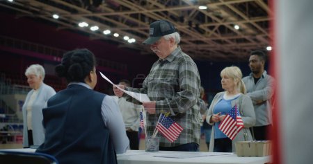 Elderly man talks with polling officer and takes vote bulletin. Multi cultural American citizens come to vote in polling station. Political races of US presidential candidates. National Election Day.