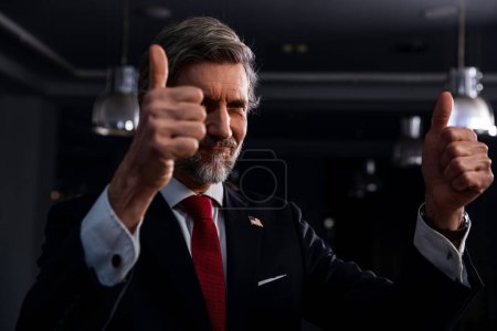 Positive mature politician comes on press conference with news journalists. Male diplomat or businessman gestures and poses for cameras for media and television live broadcast in government building.