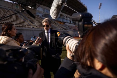 Photo for Interview of football team director at press conference for TV news. Organization representative answering questions and giving interview outside a soccer stadium. Journalists crowd on press campaign. - Royalty Free Image