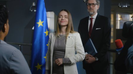 Photo for Positive female European politician smiles, poses for cameras and greets journalists. Media workers take pictures and broadcast on television. Representative of the European Union at press conference. - Royalty Free Image