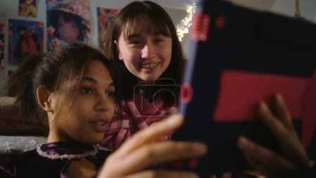 Photo for Asian teen lies on bed and watches content with friend. African American girl sits at floor, surfs internet using tablet. Multicultural girls spend leisure time together at home. Friends relationship. - Royalty Free Image