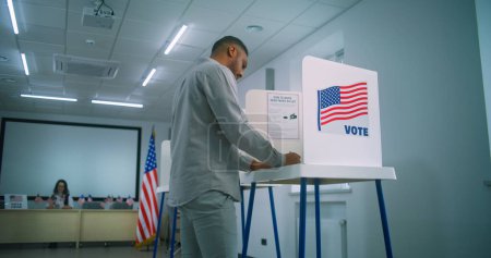 Foto de Votante masculino afroamericano camina para la mesa de registro en el centro de votación, toma boleta de papel. Cabinas de votación con el logotipo de la bandera de Estados Unidos y firma llamando para votar. Día de las Elecciones en los Estados Unidos de América. - Imagen libre de derechos