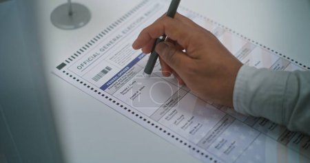 Close up of anonymous African American man choosing which presidential candidate to vote for in paper ballot in voting booth. US citizen at polling station. National Elections Day in United States.