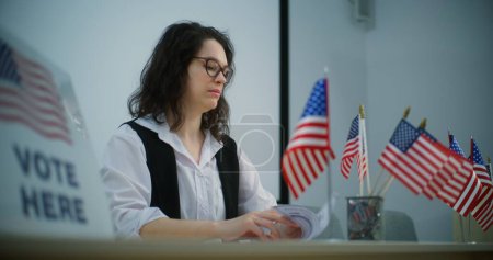 Foto de Mujer oficial de votación se sienta a la mesa con banderas estadounidenses en el centro de votación, habla con la persona sobre el registro en la votación. Día Nacional de Elecciones en los Estados Unidos. Deber cívico y democracia. - Imagen libre de derechos
