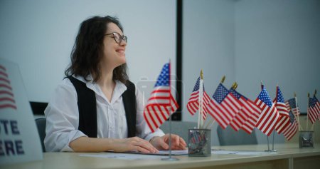 Foto de Mujer oficial de votación se sienta a la mesa con banderas estadounidenses en el centro de votación, habla con la persona sobre el registro en la votación. Día Nacional de Elecciones en los Estados Unidos. Deber cívico y democracia. - Imagen libre de derechos