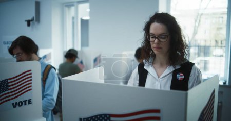 National Election Day in United States. Woman in glasses votes in booth in polling station office. Political races of US presidential candidates. Civic duty and patriotism. Slow motion. Dolly shot.
