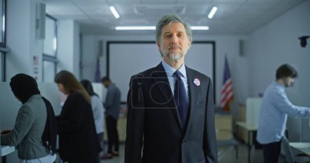Téléchargez les photos : Portrait d'un homme mûr, électeur aux États-Unis. Homme d'affaires se tient dans un bureau de vote moderne, pose et regarde la caméra, sourit. Contexte avec isoloirs et drapeaux américains. - en image libre de droit