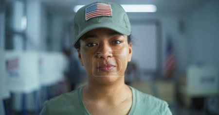 Téléchargez les photos : Portrait d'une femme soldat, électrice aux États-Unis d'Amérique. Une femme en uniforme de camouflage se tient dans un bureau de vote et regarde la caméra. Contexte avec isoloirs. Notion de devoir civique. - en image libre de droit