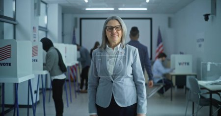 Photo for Businesswoman stands in a modern polling station, poses, looks at camera, smiles. Portrait of a mature woman, United States of America elections voter. Background with voting booths and American flag. - Royalty Free Image