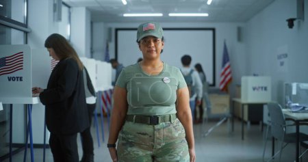 Téléchargez les photos : Une femme en uniforme de camouflage se tient dans un bureau de vote et regarde la caméra. Portrait d'une femme soldat, électrice aux États-Unis d'Amérique. Contexte avec isoloirs. Notion de devoir civique. - en image libre de droit