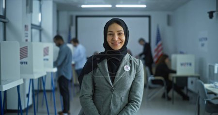 Photo for Portrait of Muslim woman, United States of America elections voter. Woman stands in a modern polling station, poses, smiles and looks at camera. Background with voting booths. Concept of civic duty. - Royalty Free Image