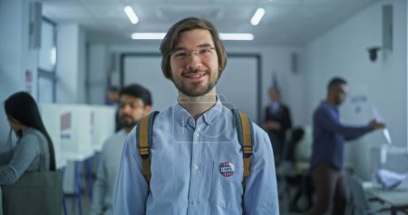 Portrait d'un homme caucasien, électeur des États-Unis d'Amérique. L'homme avec badge se tient dans un bureau de vote moderne, pose, sourit, regarde la caméra. Contexte avec isoloirs. Concept du devoir civique.